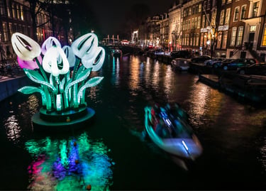 Illuminated giant tulip sculpture at the Amsterdam Light Festival on a canal with a passing tour boat.