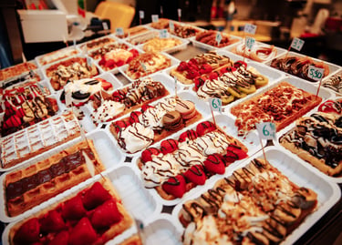 Variety of Belgian waffles with toppings like fresh strawberries, chocolate sauce, and whipped cream at a street food market.