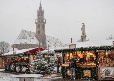 Snowy Bolzano Christmas market with wooden stalls and the Gothic cathedral spire in the background.