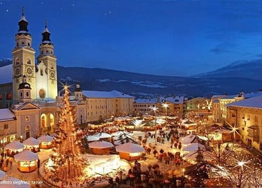 A glowing Christmas market at night in Brixen, Italy, featuring a large lit tree and the Cathedral of Bressanone.