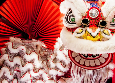 A colorful Chinese Lion Dance costume with white fur and silver scales next to a red paper fan.