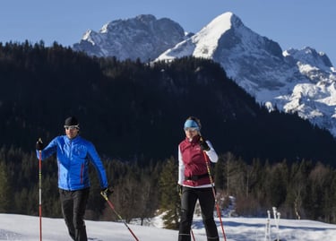 A man and woman cross-country skiing on a snowy trail with a scenic mountain range background.