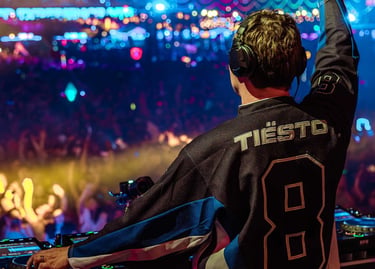 DJ Tiësto performing a live set on stage at an EDM music festival with a large cheering crowd and lights.