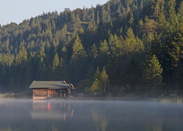 A rustic wooden boathouse on a misty mountain lake surrounded by a dense evergreen forest.