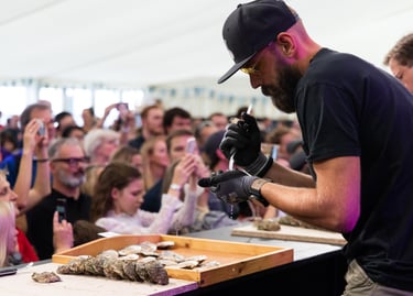 A professional chef shucking fresh oysters for a large crowd at a seafood festival demonstration.