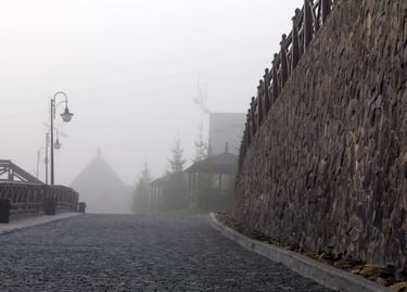 Foggy cobblestone road with a stone wall and street lamps in a misty mountain resort village.
