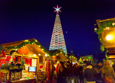 Crowds walking past illuminated wooden stalls and a giant crystal Christmas tree at an outdoor holiday market.