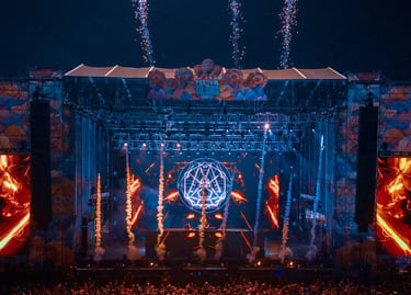 A large crowd at an outdoor music festival stage with fireworks and laser lights at night.