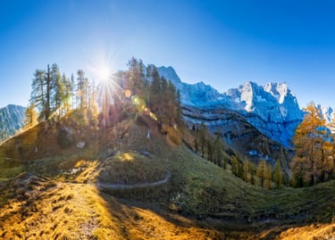Panoramic sunburst over rocky mountain peaks and golden autumn larch trees in the Alps.