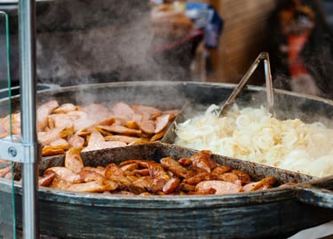 Steaming hot grilled bratwurst and sliced onions cooking in a large metal pan at a street food market.
