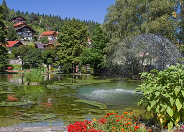 Scenic park in an Alpine village featuring a water fountain, lush gardens, and traditional mountain houses.