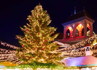 A glowing Christmas tree and festive market stalls decorated with holiday lights at night.