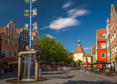 A sunny German town square featuring a traditional blue and white Maibaum maypole and historic architecture.