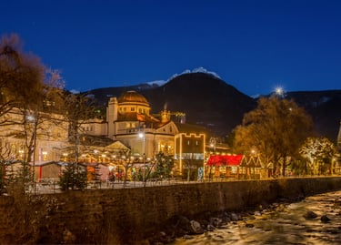 Illuminated Christmas market stalls in Merano, Italy at night with snowy mountains in the background.