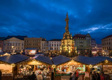 A bustling European Christmas market at dusk featuring the Holy Trinity Column in Olomouc, Czech Republic.