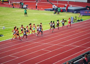 Professional female athletes competing in a long distance race on a red outdoor running track.