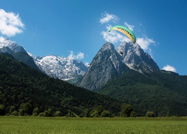 A green paraglider soaring over a lush valley and the snow-capped Julian Alps in Slovenia.