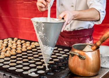 A chef uses a metal funnel to pour batter into a hot poffertjes pan to make traditional Dutch mini pancakes.