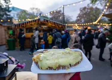 A person holding melted raclette cheese on bread at a crowded outdoor European Christmas market.