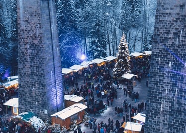 Crowded outdoor Christmas market in a snowy forest with wooden stalls and a tall decorated tree.