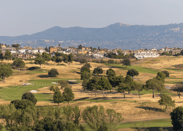 Scenic view of a lush golf course in Italy with rolling fairways, trees, and mountains in the background.