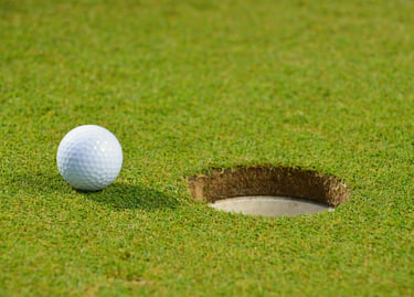 A white dimpled golf ball sits on the edge of a circular hole on a green grass golf course.