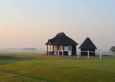 Small thatched-roof cottage and shed on a misty green golf course during sunrise.