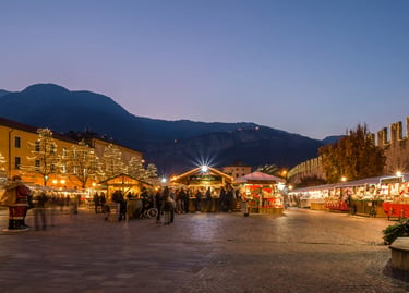 A festive European Christmas market at twilight with illuminated wooden stalls and mountains in the background.