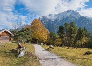 A rustic log cabin and gravel path in the Bavarian Alps with autumn trees and snow-capped peaks.