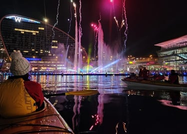 A person in a kayak watches a colorful firework and fountain show at night in Sydney's Darling Harbour.