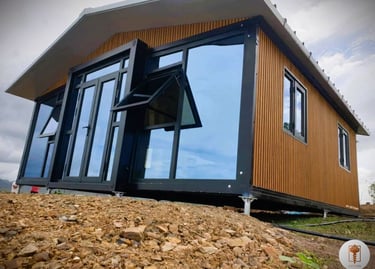 A modern prefabricated tiny home with wood siding and large glass windows on a rocky foundation.
