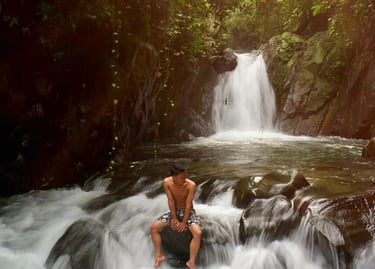 Pria bertelanjang dada duduk di atas bebatuan besar di tengah aliran air terjun deras dengan latar belakang air terjun yang l