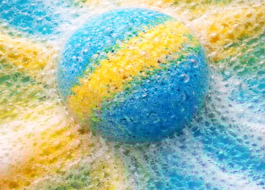 Oversized bath bomb releasing blue and yellow foam in soft, cloud-like fizz.