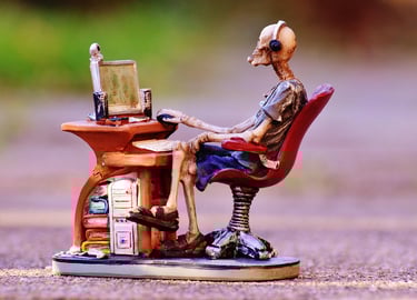 Skeleton figurine wearing a headset and sitting at a miniature computer desk gaming setup.
