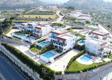 Aerial view of luxury white holiday villas with private swimming pools in a hillside Greek resort.