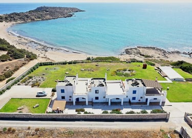 Aerial view of a white beachfront villa with blue shutters on a scenic Mediterranean coastline.