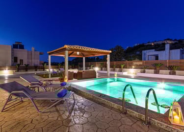 Luxury villa patio with illuminated swimming pool, lounge chairs, and a covered gazebo at night.
