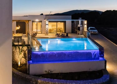 Luxury modern villa at dusk featuring an illuminated infinity pool with blue lighting and outdoor dining area.