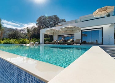 Modern luxury villa with a private infinity pool, sun loungers, and umbrellas under a clear blue sky.