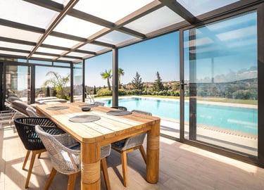 Modern covered patio with wooden dining table and glass sliding doors overlooking a luxury swimming pool.