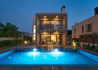 Modern luxury villa at twilight with a glowing blue swimming pool and patio lounge chairs.