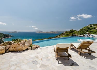 Luxury infinity pool overlooking the Aegean Sea with wooden lounge chairs on a stone terrace.