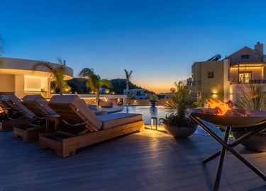 Luxury resort pool deck at sunset with wooden sun loungers, an infinity pool, and a modern fire pit.