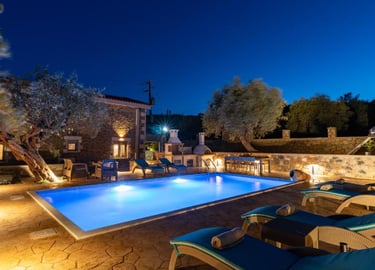 Luxury villa stone patio and private swimming pool illuminated at night with sun loungers.