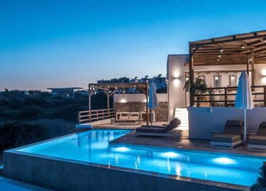 Luxury villa at twilight featuring a lit infinity pool and outdoor patio with lounge chairs.