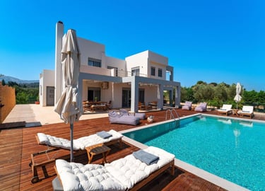 Luxury modern villa with a private swimming pool, wooden sun deck, and lounge chairs under a clear blue sky.