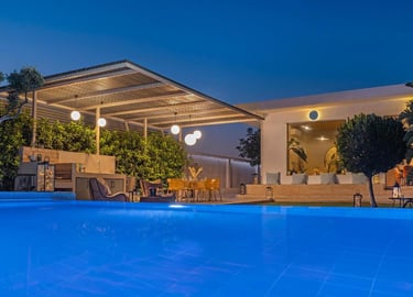 Luxury modern villa patio with blue pool, outdoor kitchen, and illuminated dining area at night.