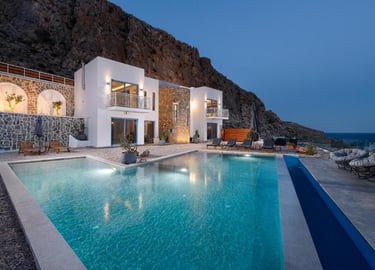 Luxury white villa with illuminated swimming pool at dusk, nestled against a rocky mountain cliff.