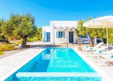 Luxury white Greek villa with a private swimming pool, sun loungers, and olive trees under a clear blue sky.