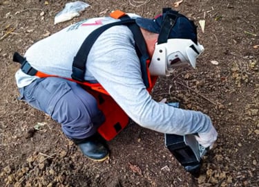 A worker in protective safety gear and face shield uses a metal detector for landmine clearance.